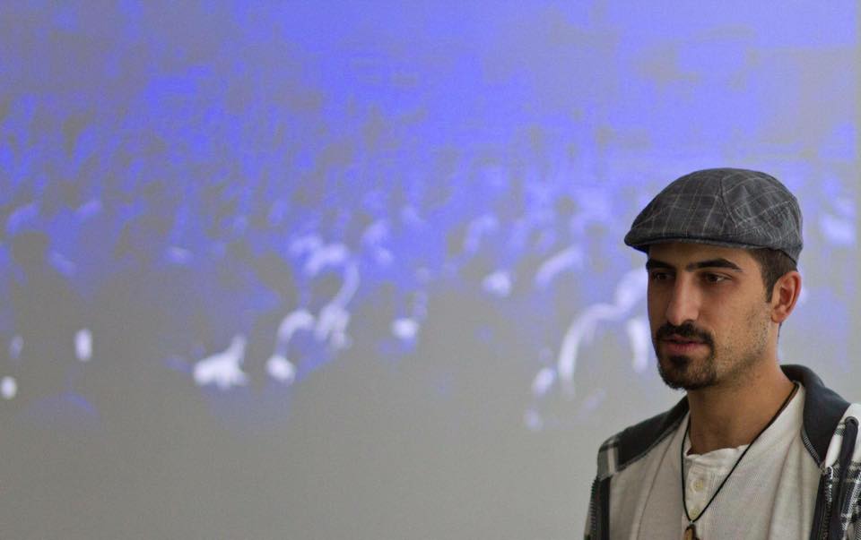 A photo of Bassel Khartabil wearing a cap standing in front of an abstract purple and gray design.