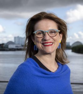 Headshot of Catherine Stihler, wearing a blue shawl standing in front of a body of water with buildings and clouds in the distance.