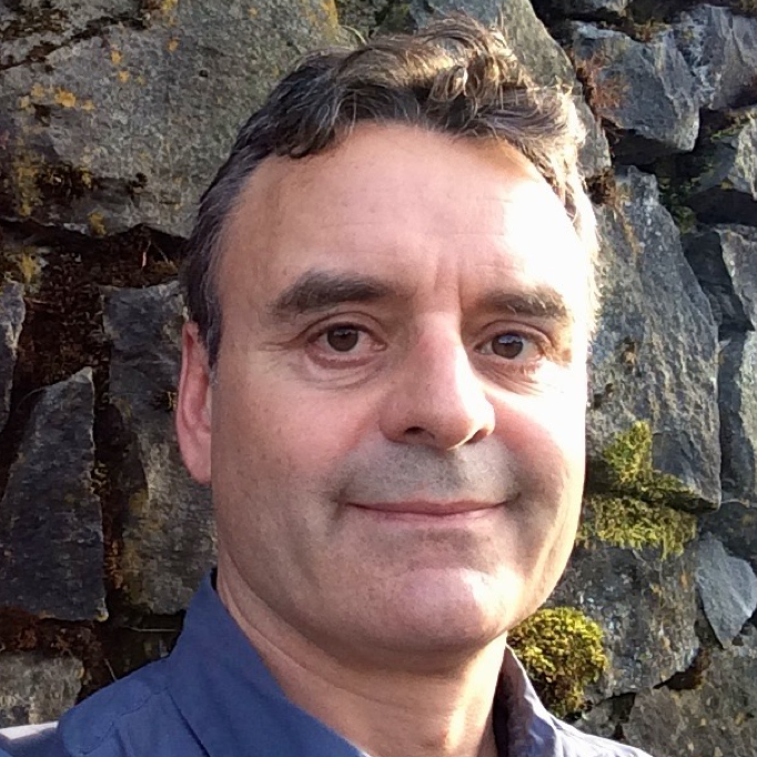 A color headshot of Nate Angell wearing a blue shirt in front of a mossy stone wall.