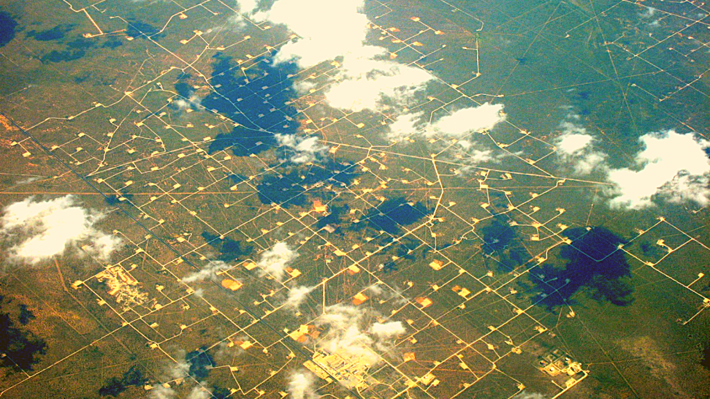 An aerial view of flat rural land with a few scattered clouds showing roads and structures that make the land look like a glowing gold circuit board.