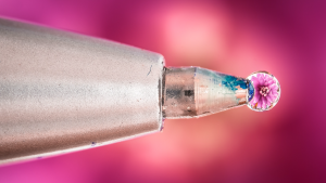 The tip of a ballpoint pen with a smear of bluish ink and a water drop reflecting/containing a pink flower blossom perched on the tip, highly magnified on a glowing pink background.