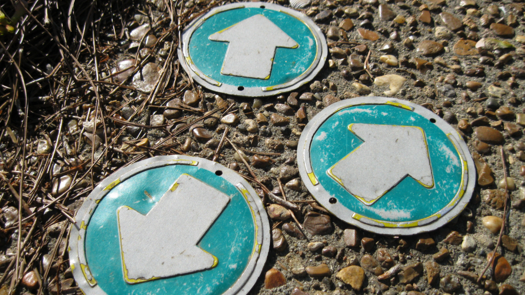 Close up photo of three round metal signs lying haphazardly on a stony path, each with a big white arrow pointing in a different direction, embossed on a greenish-blue background.
