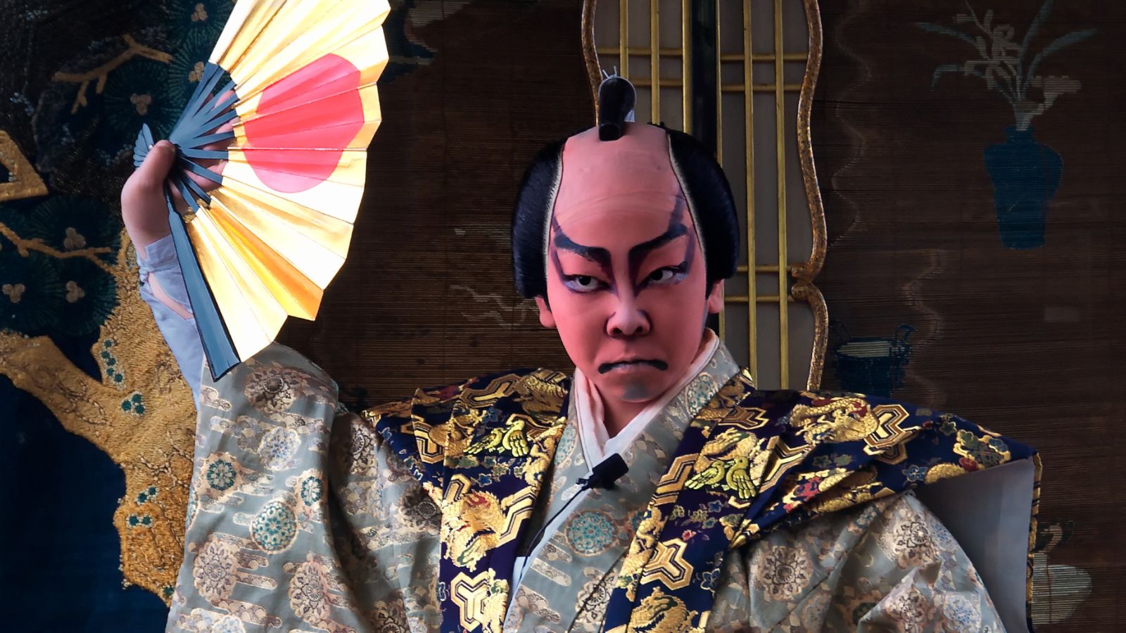 Color photo of a kabuki actor with dramatic red and black face makeup and traditional hairstyle, staring intently and wearing an ornate gold and blue costume and holding up an open yellow fan with a red dot in the middle.