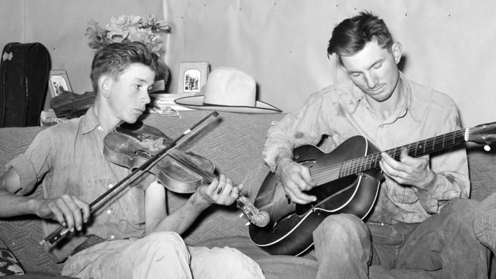 Black and white photo of two people with short, greased-back hair sitting on a couch and playing a violin and a guitar.