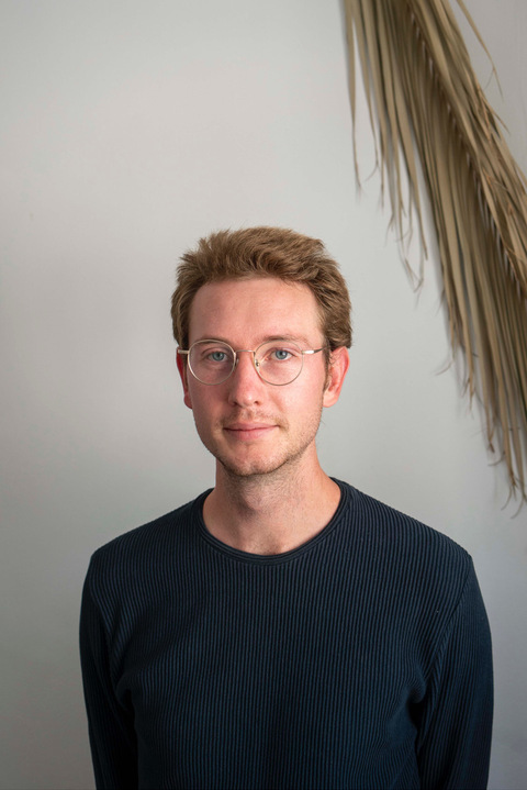 Image of Connor Benedict. Wearing glasses, with a dark shirt, in front of a palm frond.