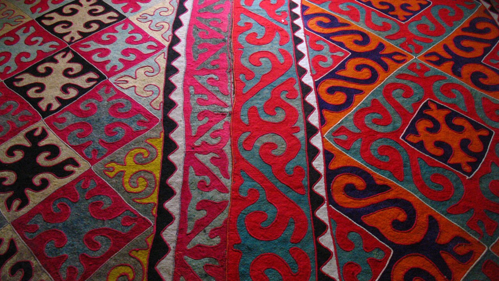 A colorful shyrdak, a type of rug, with geometric square and diamond patterns filled with spiraling and curvy abstract shapes in bright pink, red, navy, yellow, teal and light and dark blues.
