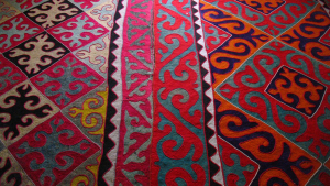 This image features a set of colorful geometric patterns of a shyrdak, a type of rug. Square and diamond patterns are filled with spiraling and curvy abstract shapes in bright pink, red, navy, yellow, teal and light and dark blues.